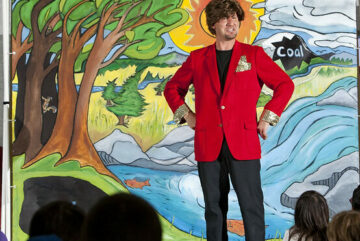 Man in red blazer and wig in front of a painted nature backdrop