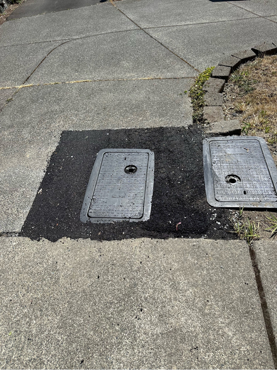 Meter box is shown in driveway with temporary asphalt patch installed.