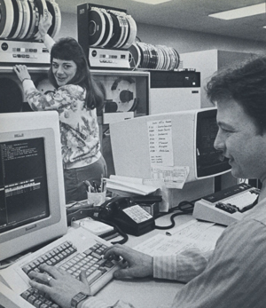 Dos trabajadores de los 80 trabajando con carretes y monitores de computadora pesados
