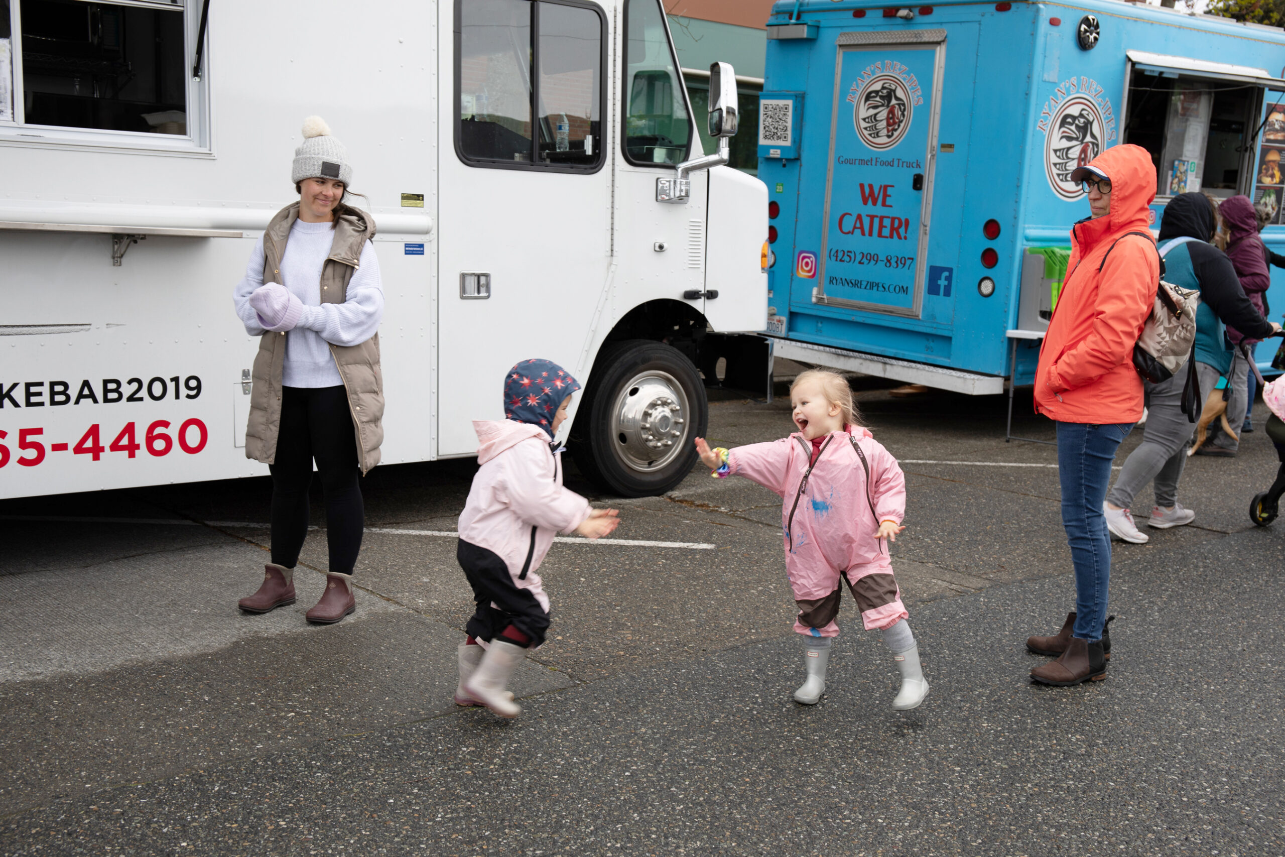 분홍색 옷을 입은 두 어린 소녀가 푸드 트럭 두 대 앞에서 쫓아다니며 놀고 있습니다.