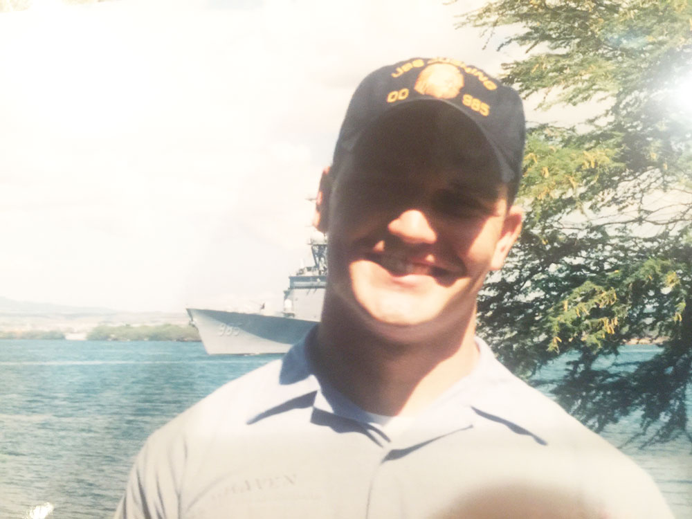 Young sailor in tropical location stands with gray US Navy ship in background