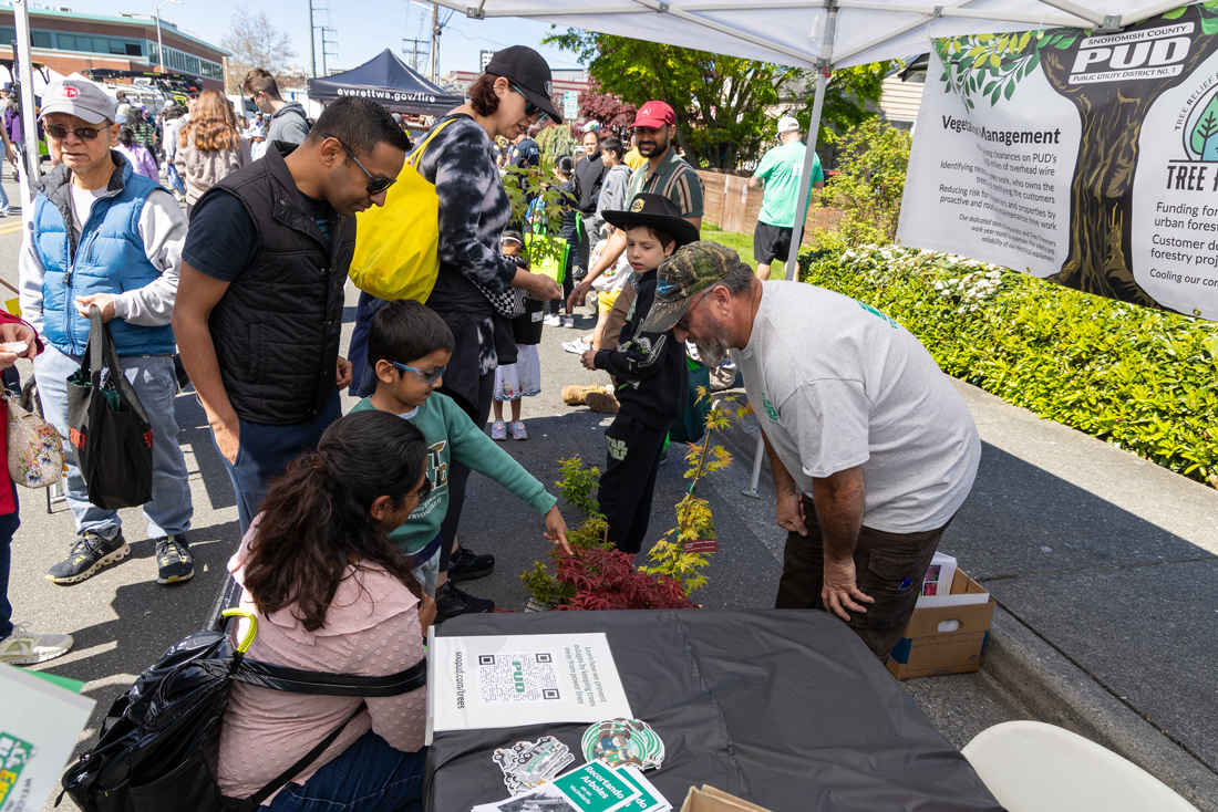 戴眼镜的小男孩指着一棵树，他的父母和几位成年人面带微笑地看着他。一位树艺师俯身靠近男孩，回答他的问题。 