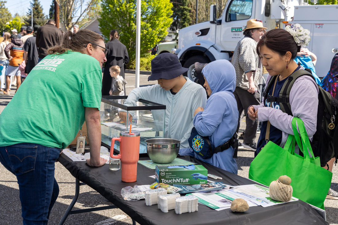 一位身穿PUD T恤的微笑女士俯身与一位母亲和她的两个孩子交谈，他们正在户外桌旁进行一项教育实验。