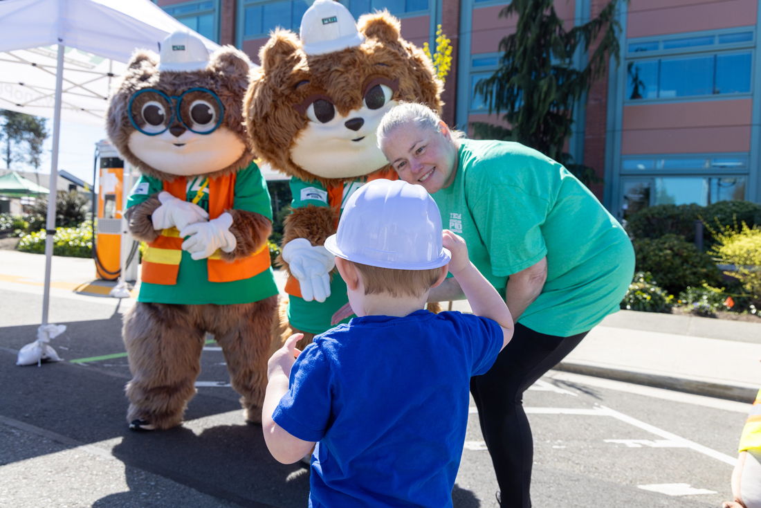 一位女士面带微笑，俯身和一个小孩说话。小孩戴着玩具安全帽，害羞地看着PUD的吉祥物Zip和Zap。