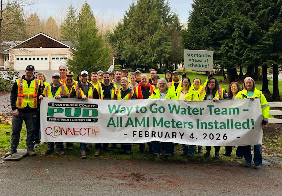 PUD Water Team celebrates early completion of new water meter installs. Large group of people in utility safety gear holding a banner.