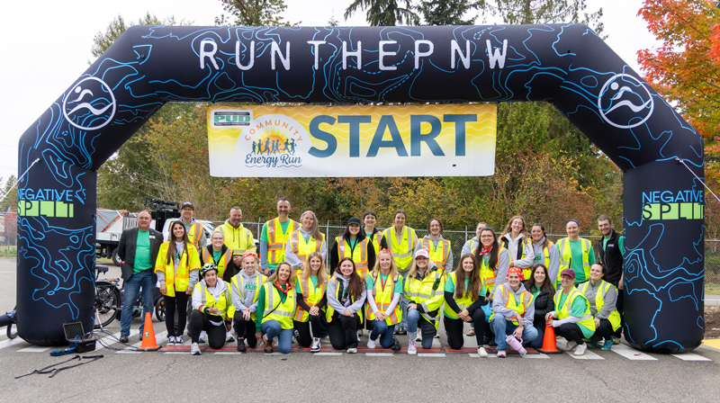 Community Energy Run volunteers and staff wearing high-visibility safety vests pose for a group photo beneath the starting arch at the event start line.