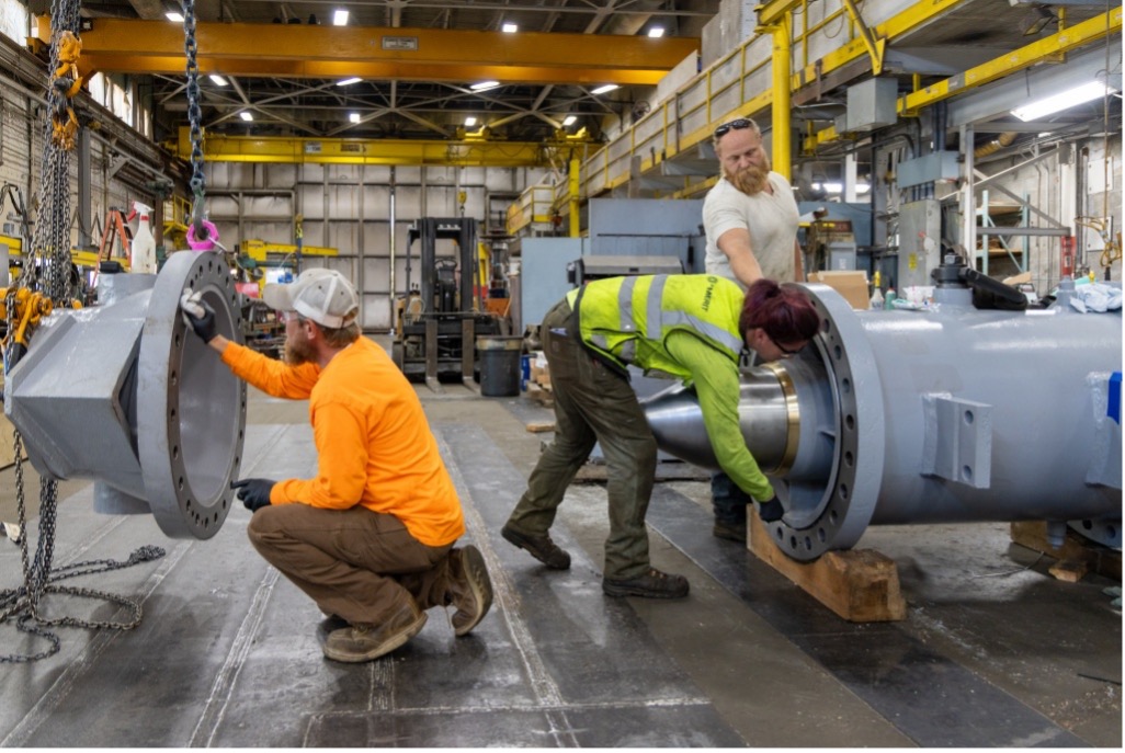 Three PUD employees work together inside a maintenance shop, aligning and assembling a large industrial metal component using lifting equipment and hand tools.