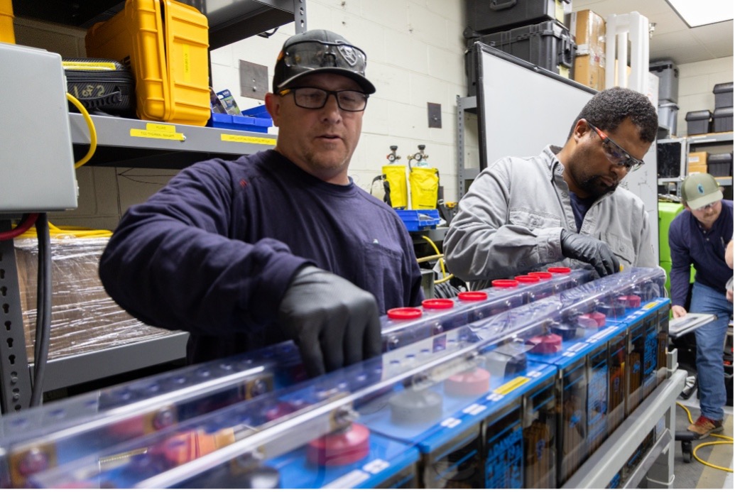 PUD employees install and inspect electrical equipment, working at a rack in a workshop environment.